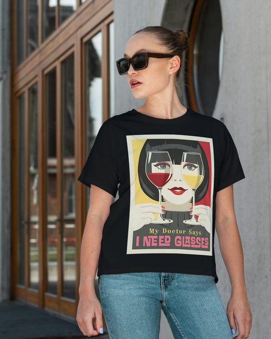 A black short-sleeve crewneck t-shirt featuring a retro-style graphic print of a woman with glasses on her face, holding wine glasses, with the text 'My Doctor Says I Need Glasses'. The t-shirt is displayed flat against a neutral light gray background.